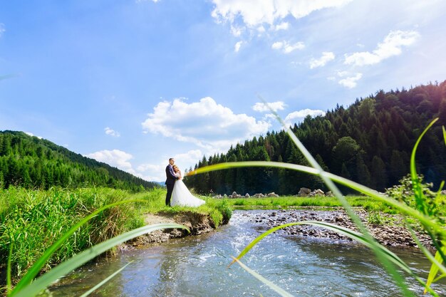 Sposi innamorati in abito bianco e vestito a piedi in estate vicino al fiume.