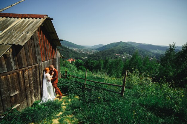 Wedding couple in the mountains. The groom and bride are hugging near the rustic house. A beautiful view of the mountains behind the couple