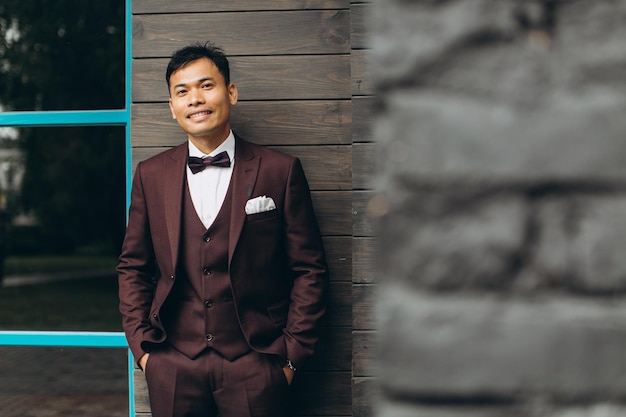Wedding day Asian groom poses on the background of a wooden building and large windows