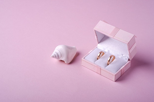 Wedding gold rings in pink gift box with white seashell on soft pink background, angle view, copy space