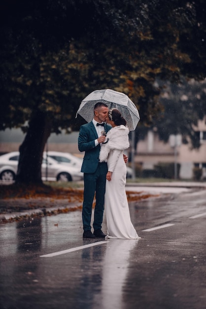 結婚式の幸せな瞬間。幸せな若いカップル、傘をさしながら雨天の通りを歩く新婚夫婦。高品質の写真