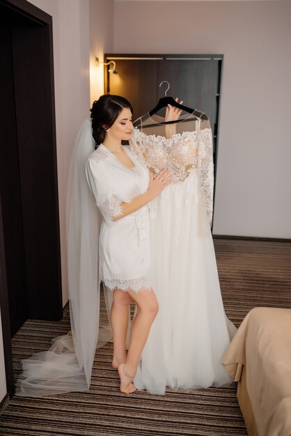 Wedding morning preparation. Happy bride holds in her arms and looks at her wedding dress.