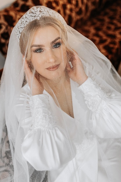 Wedding portrait Blonde bride in a silk suit and tiara poses behind a sheer veil while sitting in a chair in her room