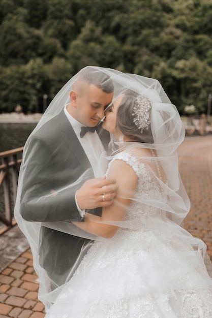 Foto ritratto di matrimonio una sposa bruna in un vestito lungo e uno sposo in un abito classico posano sotto un velo e la baciano sulla guancia