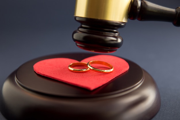 木の傷ついた心の形に結婚指輪、木の背景に裁判官のハンマー。離婚訴訟