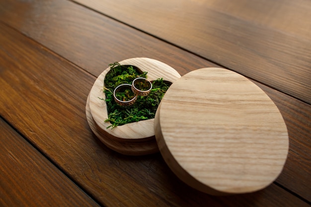 Wedding rings in ring box in shape of heart with green moss on wooden background with copy space. Rustic wedding concept.