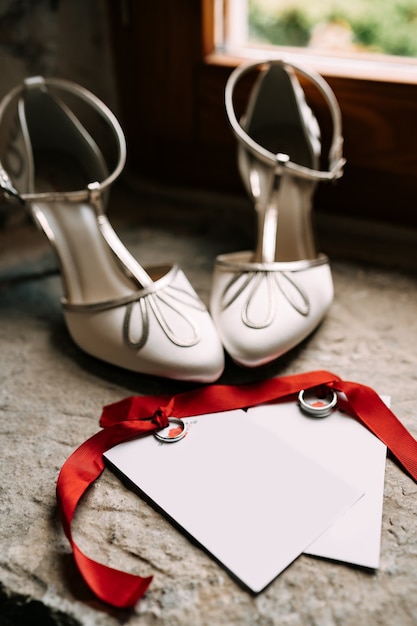 빨간 리본으로 묶인 결혼 반지는 흰색 하이힐 옆에 장식품이있는 빈 엽서에 누워 있습니다.