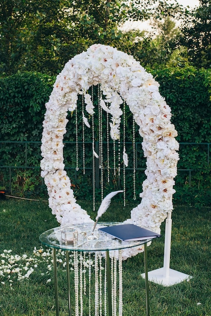Wedding rings with a glass jewelry box next to a pen for writing on a glass table decorated with glass beads against the background of a white flower arch