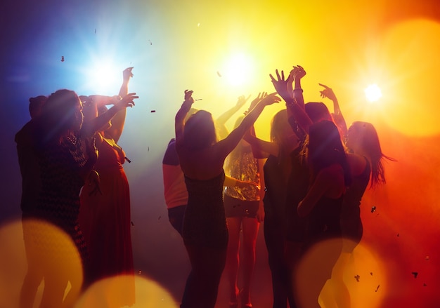 Фото Выходные. толпа людей в силуэте поднимает руки на танцполе на фоне неонового света. ночная жизнь, клуб, музыка, танцы, движение, молодежь. желто-голубые цвета и подвижные девочки и мальчики.