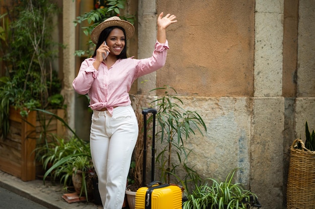週末は、帽子をかぶった幸せなスタイリッシュな若いミレニアル世代の黒人女性観光客を、スーツケースを持ってつなぐ