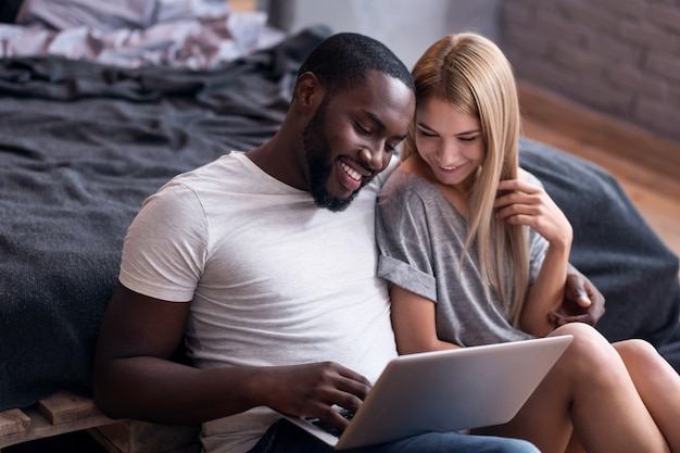 Weekend is fun. Joyful young international couple sitting in bedroom and using laptop while spending time together.