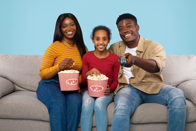 Weekend Leisure Black Family Watching Tv And Eating Popcorn Sitting On Couch Over Blue Studio Background