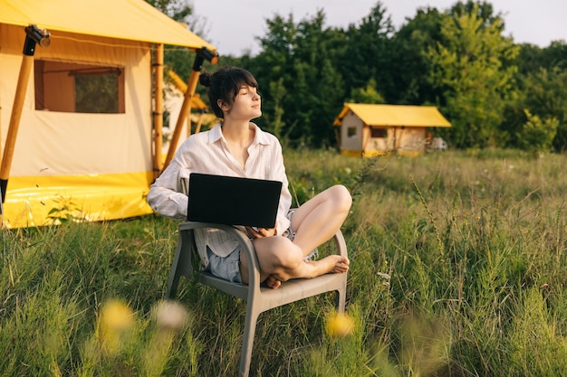 Weekend work Woman starts her morning working on a laptop while sitting near a tent in a glamping site for tourists