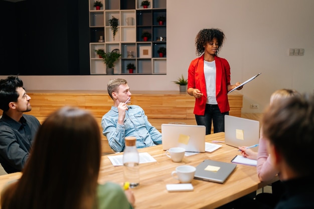 Weergave op afstand van een divers zakelijk team in de directiekamer, luisterend naar een vrouwelijke Afro-Amerikaanse manager die verslag uitbrengt over de voortgang van het project