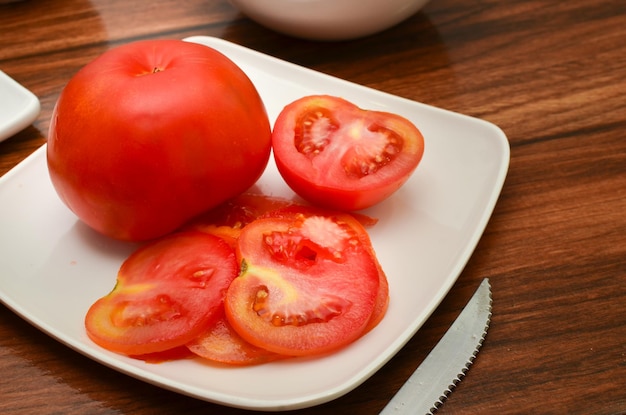 Weergave van een hele rode tomaat en een gesneden op een witte plaat met een mes naast de plaat op de houten tafel Diagonale weergave van bovenaf