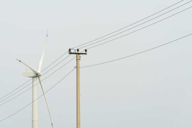 Weergave van een moderne windmolen tegen een blauwe lucht De witte bladen van de windturbine Hernieuwbare energiebron Oude hoogspanningslijnen naast moderne windturbines