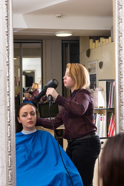 Weerspiegeling in de spiegel van blonde stylist die föhn gebruikt om haar te drogen van vrouwelijke brunette cliënt die een kiel draagt en in de stoel in de salon zit