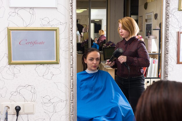 Weerspiegeling in spiegel van jonge brunette vrouw met nat haar gedroogd door blonde stylist met behulp van hete lucht föhn in salon, ingelijst door certificaat van onderwijs referenties opknoping op muur