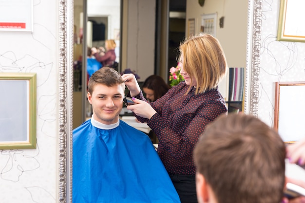 Weerspiegeling in spiegel van lachende jonge man die haar heeft geknipt en gestyled door blonde vrouwelijke stylist in salon