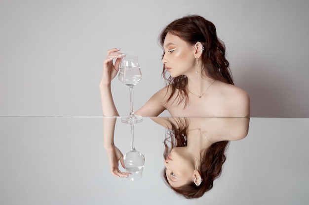 Weerspiegeling van een vrouw in spiegel met glas in haar hand. Mooie gezichtsmake-up, natuurlijke cosmetica