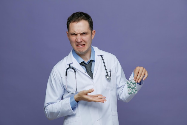 Weerzinwekkende jonge mannelijke arts met een medisch gewaad en een stethoscoop om de nek met een pak capsules die de hand erop richten en naar de camera kijken die op een paarse achtergrond wordt geïsoleerd