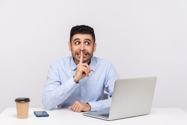 Wees alsjeblieft stil Grappige positieve man werknemer zit kantoor werkplek met laptop vragen om stilte of geheimhouding vinger op de lippen houden en vertrouwelijke informatie studio shot geïsoleerd stilzwijgen