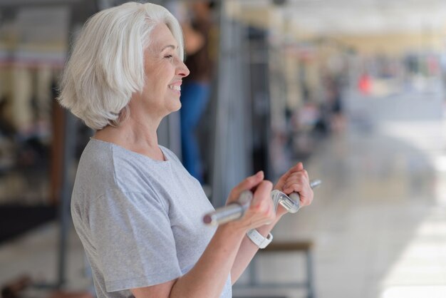 Wees gezond. Tevreden vrij senior vrouw barbell opheffen en glimlachen tijdens het sporten in een sportschool.