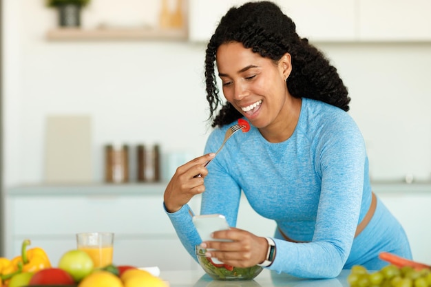 Weight loss app happy black lady using smartphone while eating\
vegetable salad in kitchen at home copy space