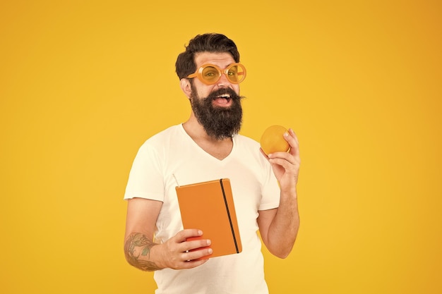 Weight loss and nutrition literature bearded man holding orange fruit and book for literature reading on yellow background dieting literature information literature on healthy diet and nutrition