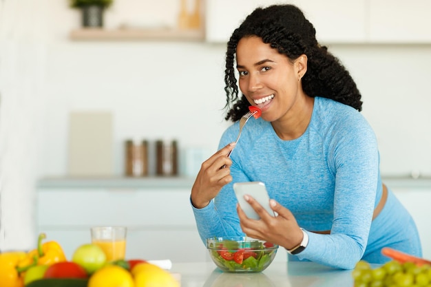 Photo weightloss app african american sporty lady using cellphone and eating fresh salad having dinner after workout