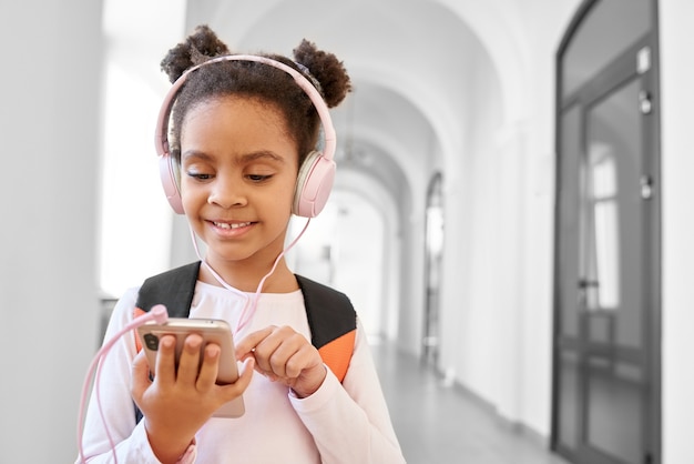 Weinig afrikaans schoolmeisje die oproeptelefoon gebruiken en muziek van roze hoofdtelefoons luisteren.