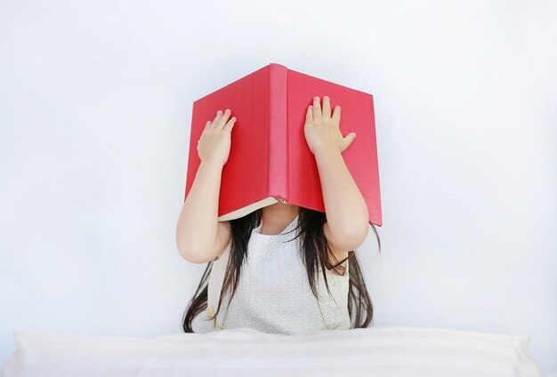 Weinig Aziatisch kindmeisje die gezicht met open hardcover boekzitting op bed met hoofdkussen verbergen.