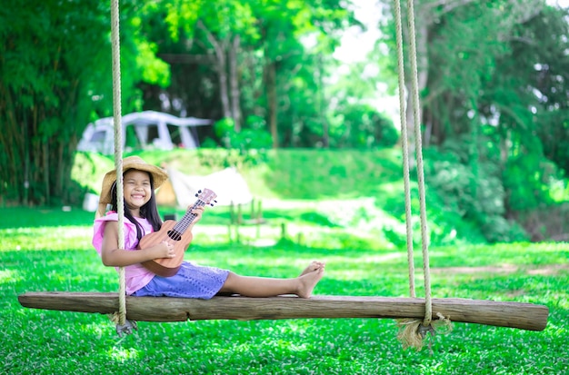 Weinig Aziatisch meisje zittend op houten schommel ukulele spelen tijdens het kamperen in natuurpark