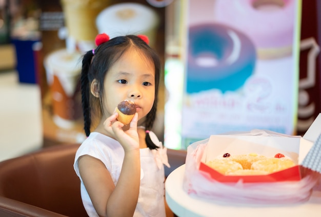 Weinig Aziatische meisjeszitting op de stoel met het houden en het eten van doughnuts