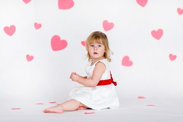 Weinig blondemeisje in witte kleding met rood lint op witte vloer met roze harten op de Valentijnskaartendag