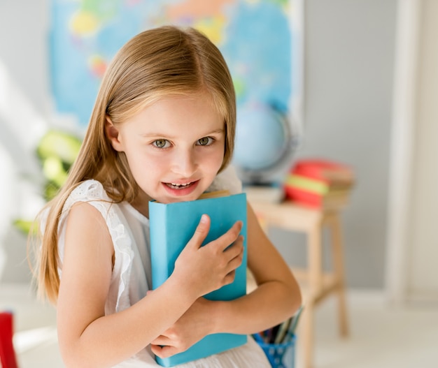 Weinig glimlachend blond meisje dat blauw boek in de schoolklasse houdt