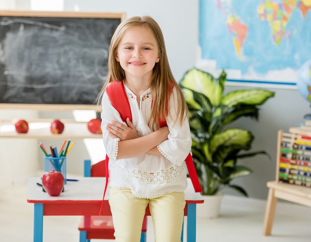 Weinig glimlachend blond meisje dat zich dichtbij bureau met haar rode zak in de ruime schoolklasse bevindt