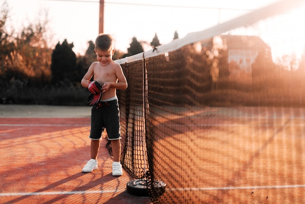 Weinig jongen die zich op tennisspeelplaats bevindt en handschoenen op zijn handen zet. Hij bereidt zich voor op training buiten.