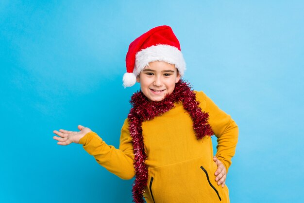 Weinig jongen het vieren Kerstmisdag die een geïsoleerde santahoed draagt tonend een exemplaarruimte op een palm en het houden van een andere hand op taille.