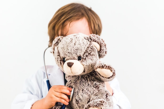 Foto weinig jongen kleedde zich als arts die teddybeer op witte achtergrond ausculteert