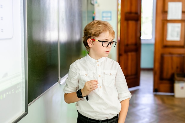 Weinig jongen met grote zwarte glazen en wit overhemd die zich dichtbij schoolbord bevinden met een krijtje die slim het denken gezicht maken