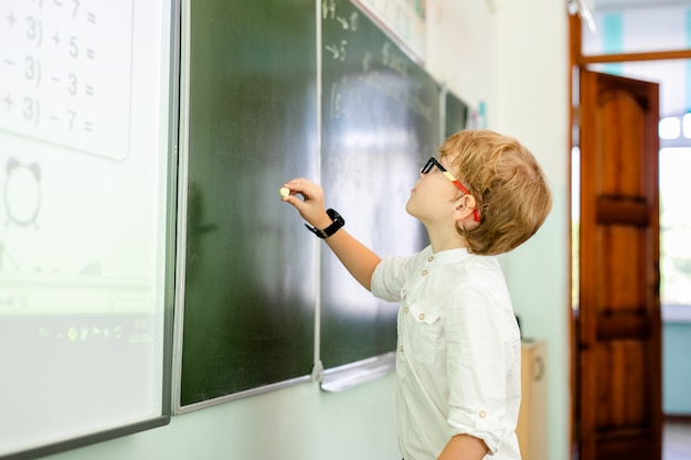 Weinig jongen met grote zwarte glazen en wit overhemd die zich dichtbij schoolbord bevinden met een krijtje die slim het denken gezicht maken