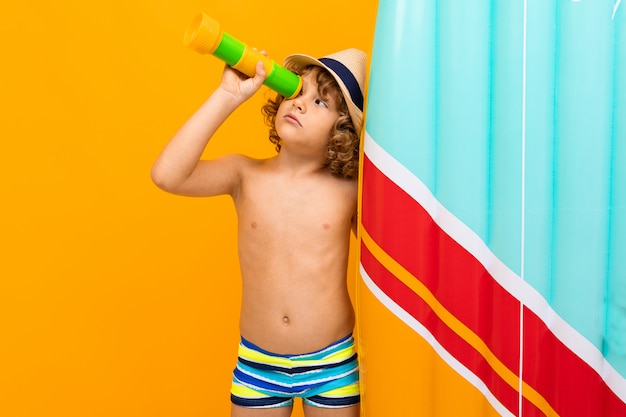 Weinig jongen met krullend haar in zwempak met rubbermatras dat op geel wordt geïsoleerd