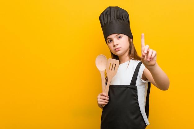 Weinig kaukasisch meisje die een chef-kokkostuum dragen die nummer één met vinger tonen.