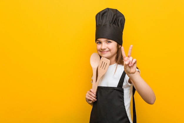 Weinig Kaukasisch meisje die een chef-kokkostuum dragen die nummer twee met vingers tonen.