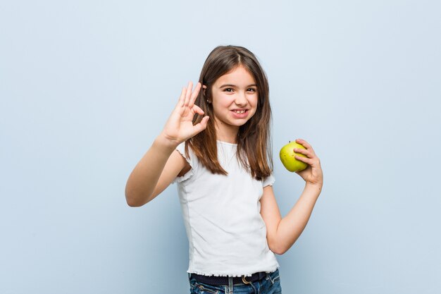 Weinig Kaukasisch meisje die een groene appel houden vrolijk en zeker tonend ok gebaar.