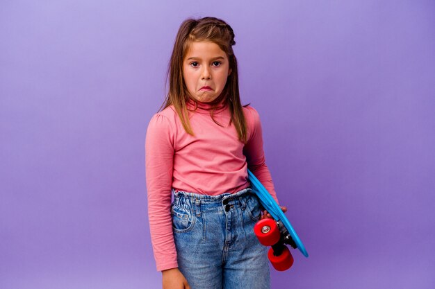 Weinig Kaukasisch meisje van de skater dat op blauwe muur wordt geïsoleerd haalt schouders op en opent verwarde ogen.