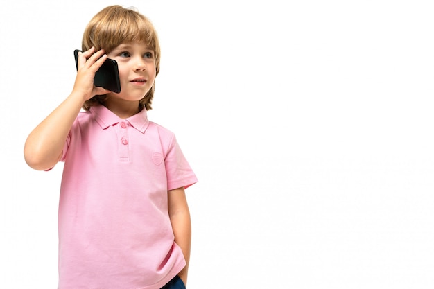 Weinig Kaukasische jongen met roze t-shirt roept een telefoon die op witte achtergrond wordt geïsoleerd