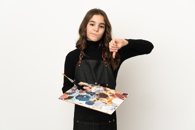 Weinig kunstenaarsmeisje die een palet houden dat op witte achtergrond wordt geïsoleerd die duim met negatieve uitdrukking toont