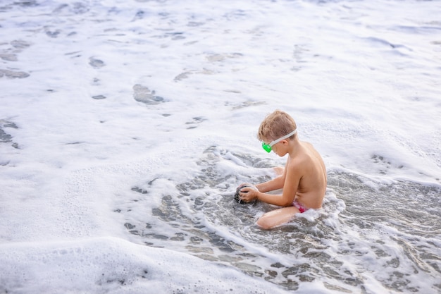 Weinig leuke Kaukasische peuterjongen met blond haar speelt bij het strand in overzeese golven op zonnige de zomerdag.